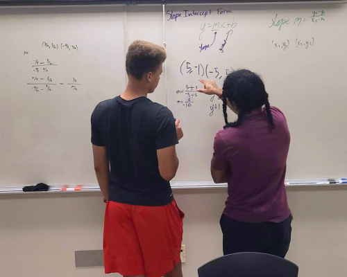 The Academy SPS student, Keon Lewis, is doing a math problem on the board and receiving help from the teacher.