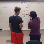 The Academy SPS student, Keon Lewis, is doing a math problem on the board and receiving help from the teacher.